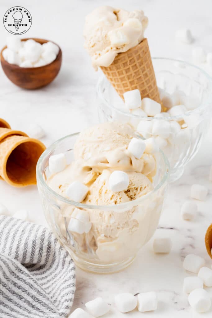 a glass mug filled with marshmallow ice cream topped with mini marshmallows. Behind that is another glass mug with a cone filled with ice cream.