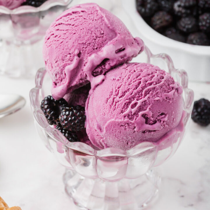 three scoops of purple, black raspberry ice cream in a scalloped glass ice cream dish with black raspberries for garnish.