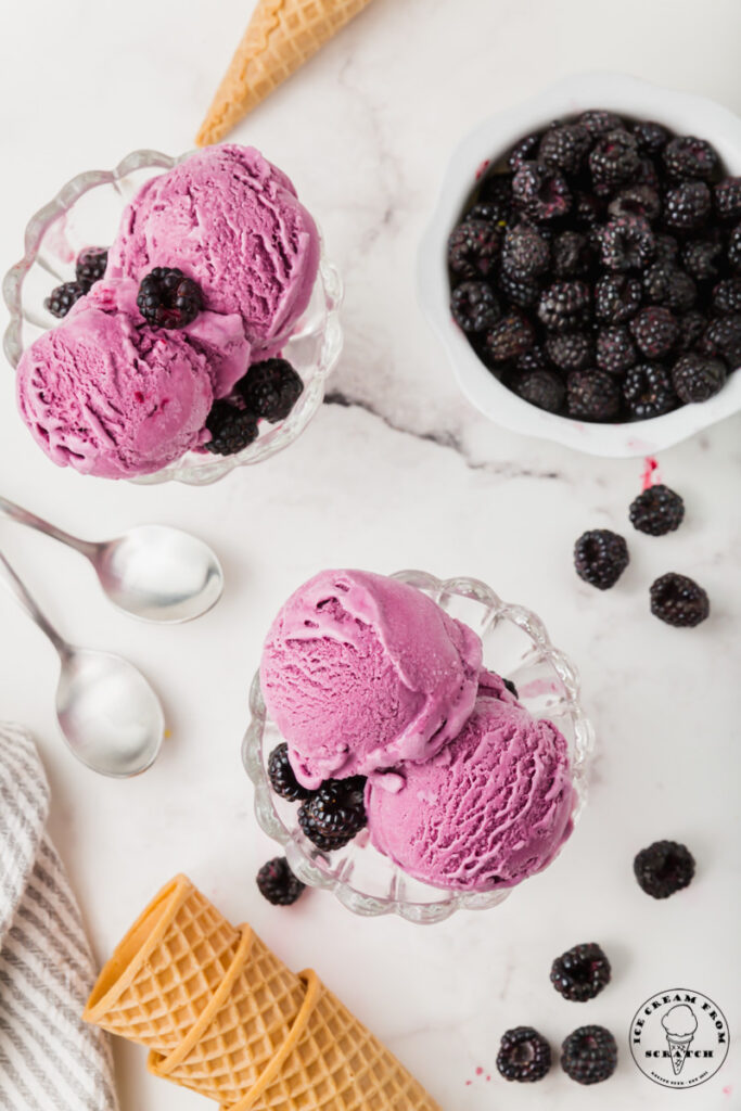 top down image of a marble countertop with two glass dishes of black raspberry ice cream, a white bowl of dark raspberries, two spoons, and sugar cones.