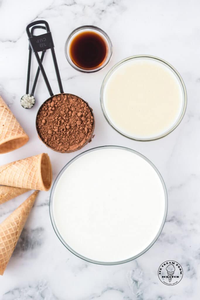 Ingredients for no churn chocolate ice cream in separate bowls on a marble counter, along with sugar cones.