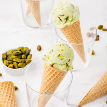 three glasses, two with cones of pistachio ice cream in them. One with a stack of sugar cones. Glasses are surrounded by pistachios