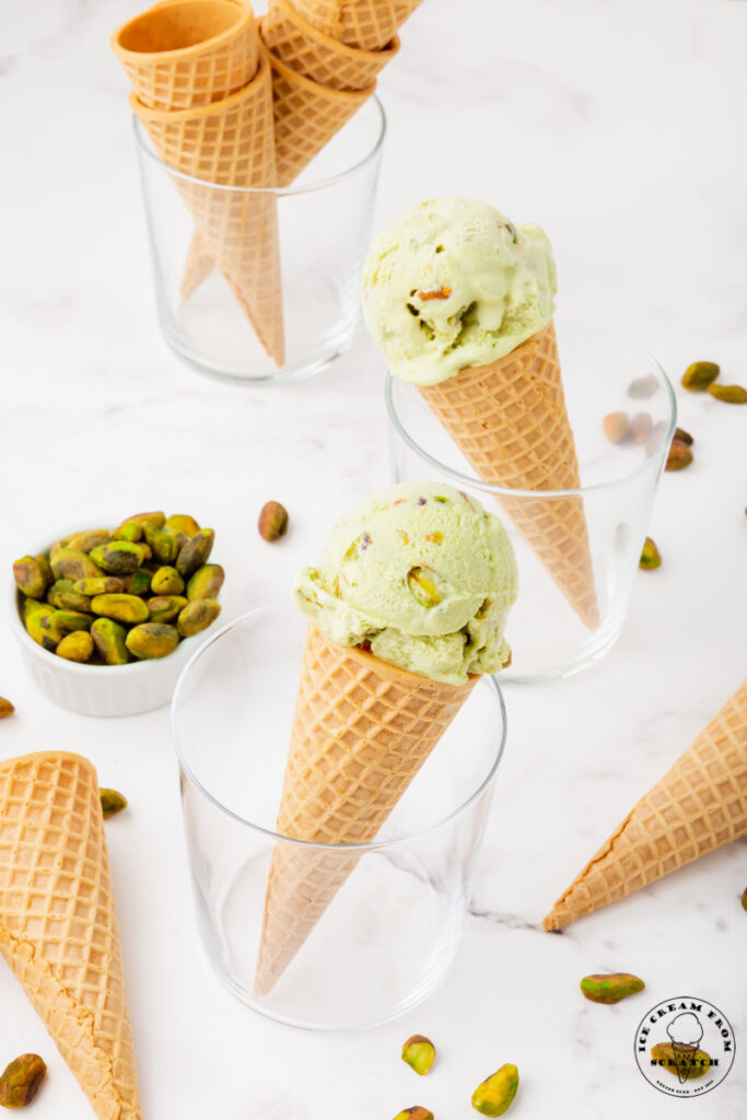 three glasses, two with cones of pistachio ice cream in them. One with a stack of sugar cones. Glasses are surrounded by pistachios