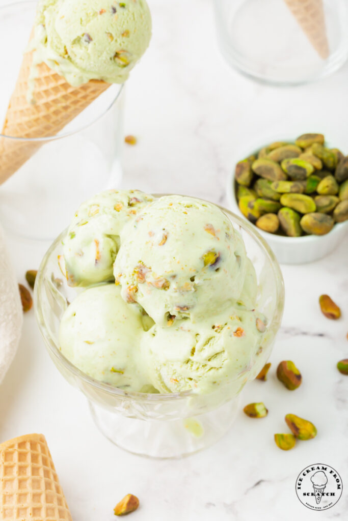 a glass ice cream dish with multiple scoops of pistachio ice cream. In the background is a small bowl of pistachios and a sugar cone filled with pistachio ice cream.