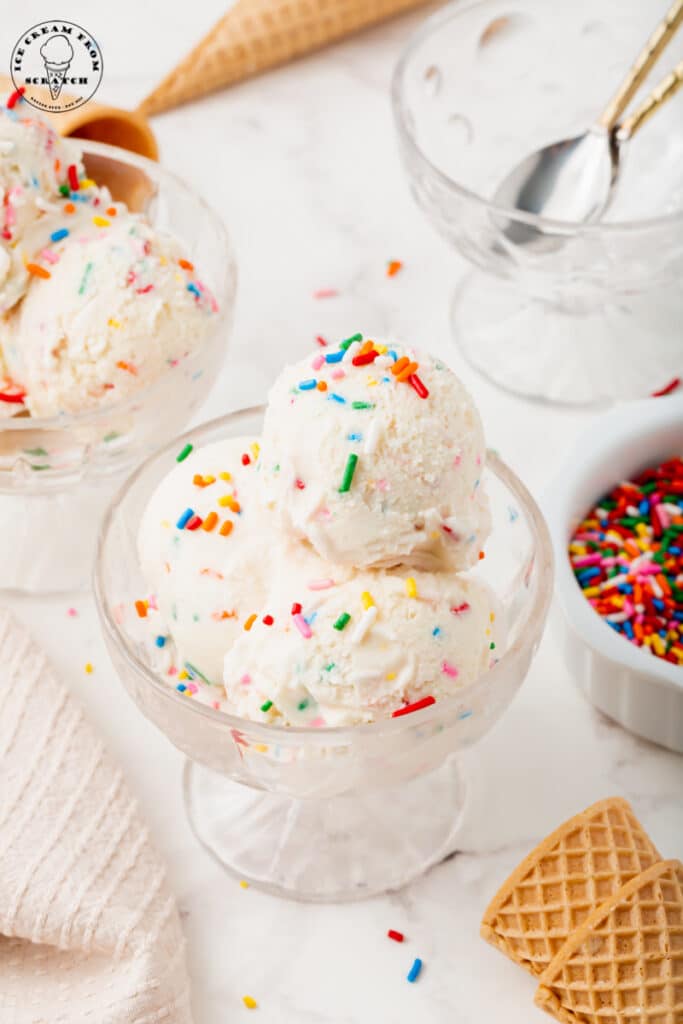 a glass dish with three scoops of rainbow sprinkle ice cream, surrounded by more sprinkles, ice cream cones, and a dish with spoons.