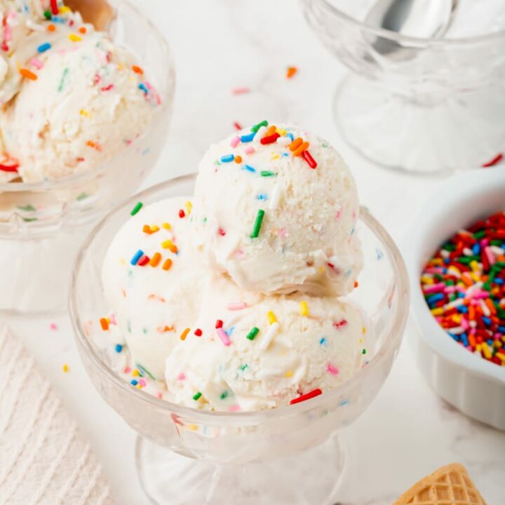a glass dish with three scoops of rainbow sprinkle ice cream, surrounded by more sprinkles, ice cream cones, and a dish with spoons.