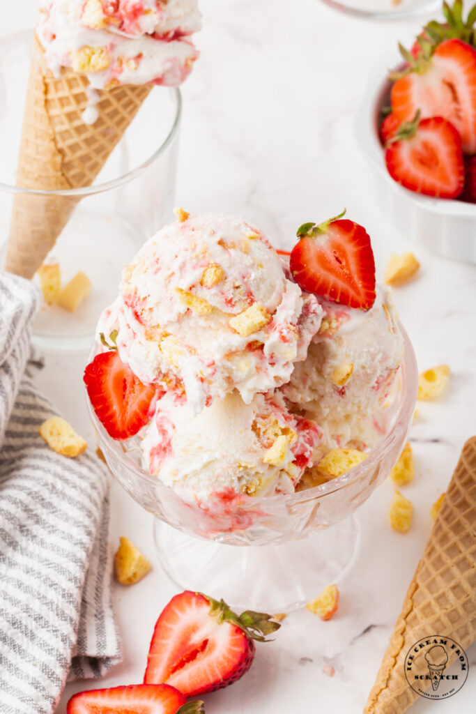 Strawberry shortcake ice cream garnished with fresh strawberries in a glass ice cream dish. In the background is a cone of strawberry ice cream and a bowl of strawberries.