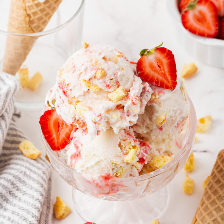 Strawberry shortcake ice cream garnished with fresh strawberries in a glass ice cream dish. In the background is a cone of strawberry ice cream and a bowl of strawberries.