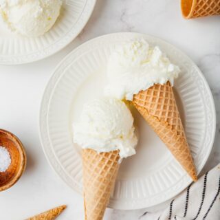 two sugar cones of white, sweet cream ice cream set on a white scalloped plate.