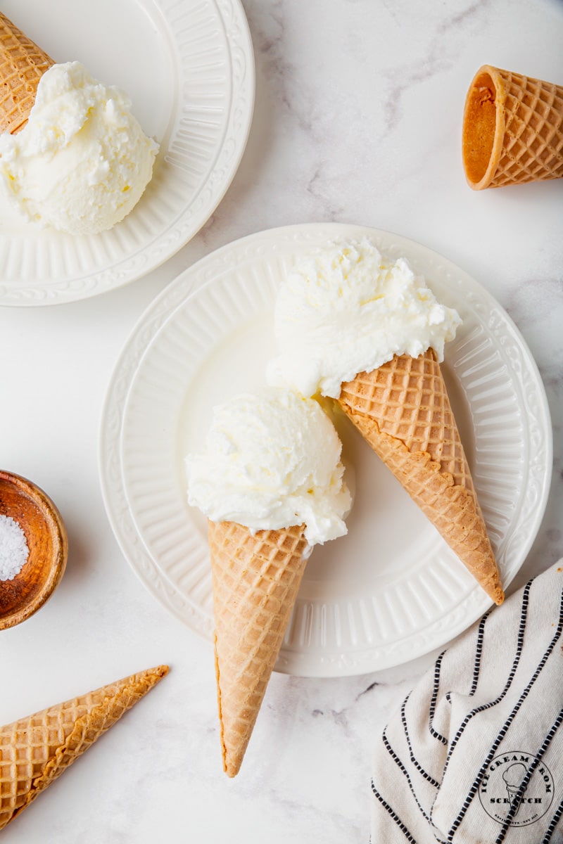 two sugar cones of white, sweet cream ice cream set on a white scalloped plate.