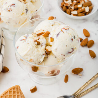 two glass ice cream dishes filled with toasted almond ice cream, surrounded by almonds, sugar cones, and two spoons.