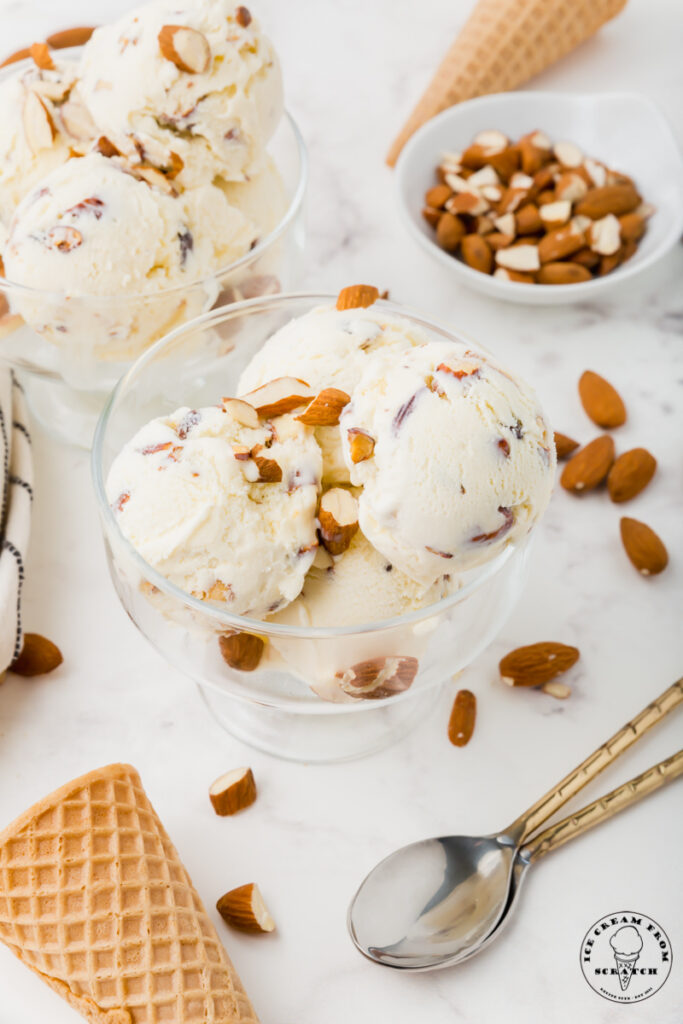 two glass ice cream dishes filled with toasted almond ice cream, surrounded by almonds, sugar cones, and two spoons.