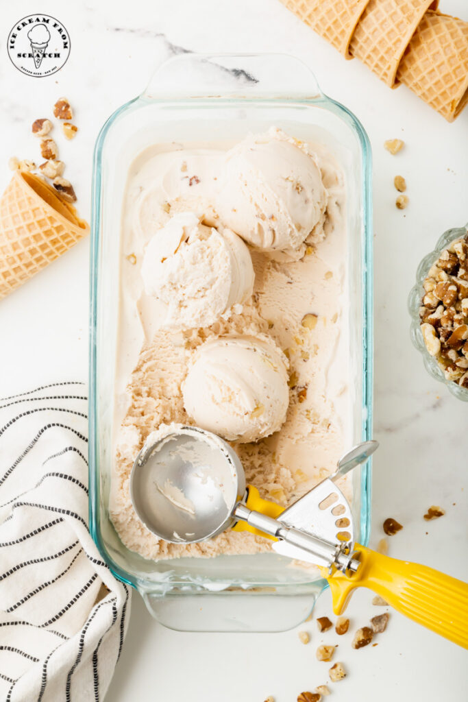 a glass loaf pan with handles, filled with black walnut ice cream. Three scoops have been made with a yellow handled metal ice cream scoop. Around the dish are sugar cones and chopped nuts.