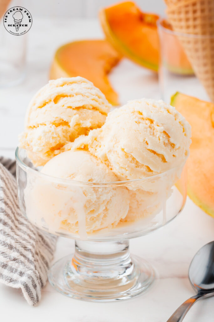 four scoops of melon ice cream in a glass ice cream dish. There are slices of cantaloupe in the background.