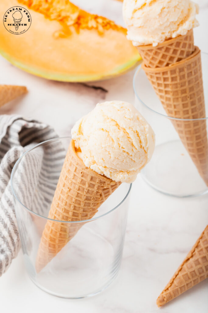 a sugar cone with a scoop of melon ice cream, propped up in a glass in front of another melon ice cream cone in another glass. A wedge of orange cantaloupe is in the background