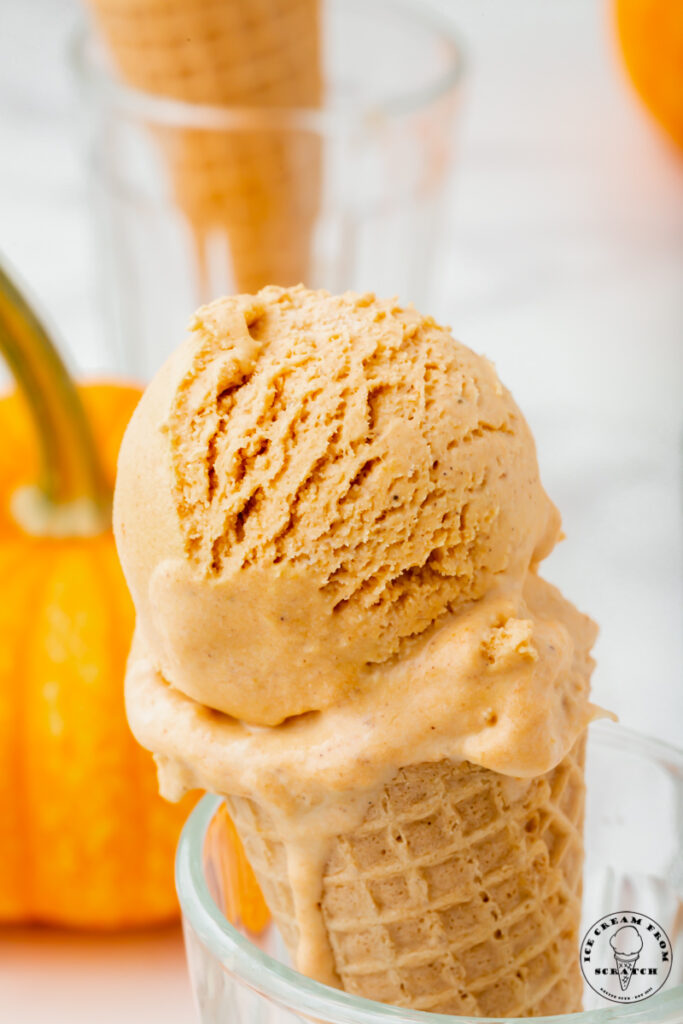 a sugar cone with a scoop of pumpkin spice ice cream, propped up in a glass. There is a pumpkin in the background.