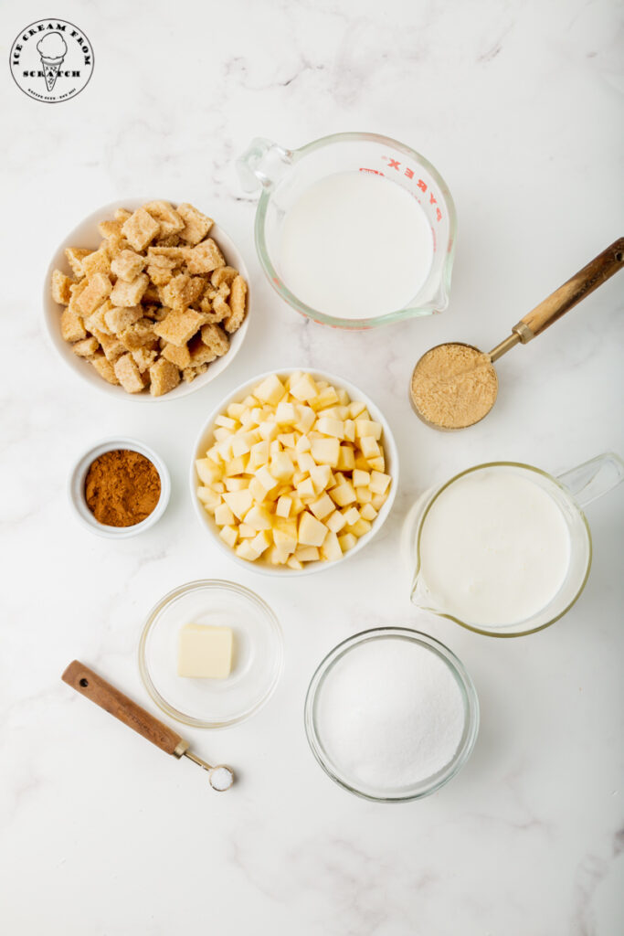 Ingredients for apple ice cream, each in separate bowls on a marble countertop. Including cookies, apples, cinnamon, and dairy items.