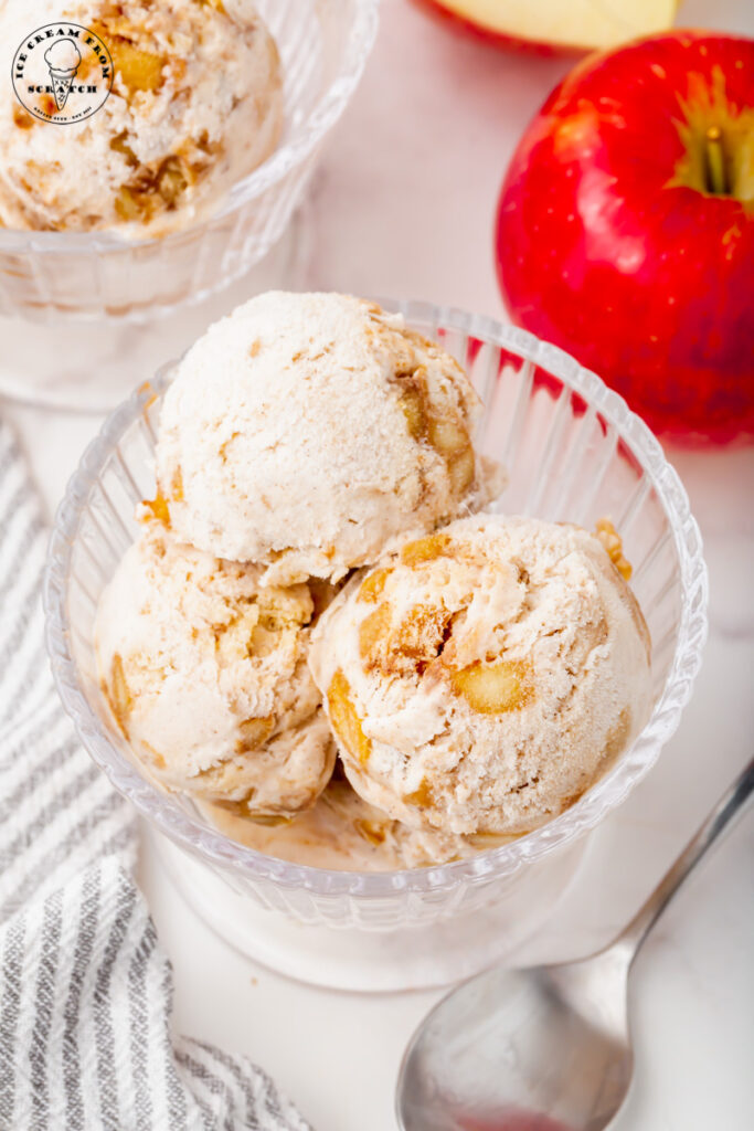 overhead shot of a glass ice cream dish with three scoops of homemade apple pie ice cream. Next to the dish is a silver spoon and a red apple