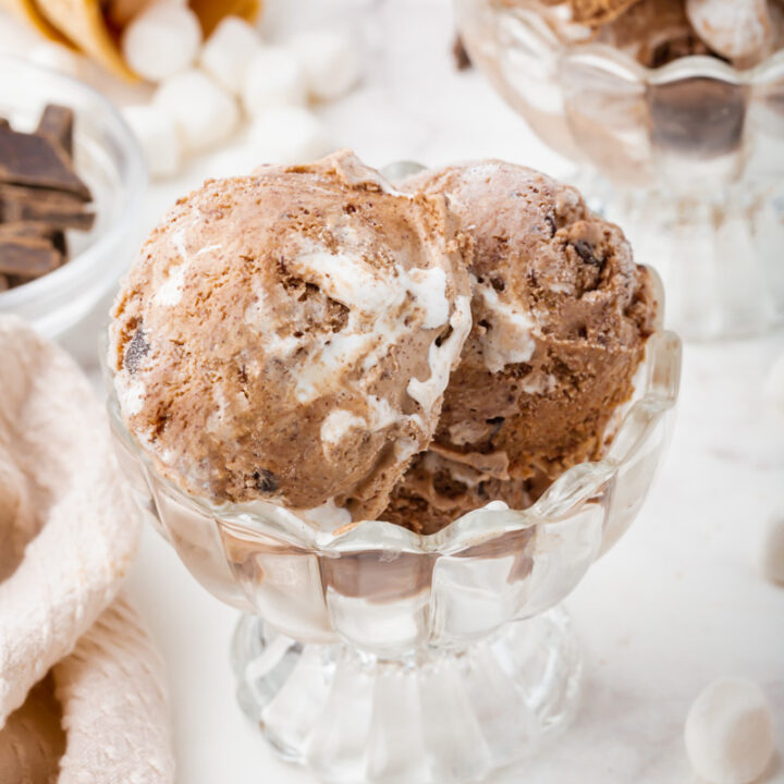 Two scopos of chocolate marshmallow ice cream in a glass dish. Mini marshmallows are strewn around as well as ice cream cones and chocolate chunks.