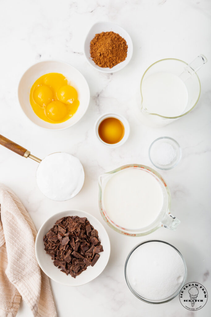 Ingredients for homemade chocolate marshmallow ice cream, each in separate bowls on a marble countertop. Includes egg yolks, cocoa powser, sugar, chocolate, and dairy items.