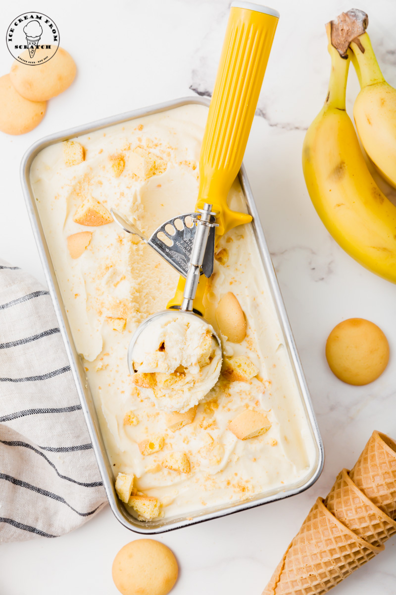 a metal pan filled with banana ice cream topped with vanilla wafers being scooped with a yellow ice cream scoop.