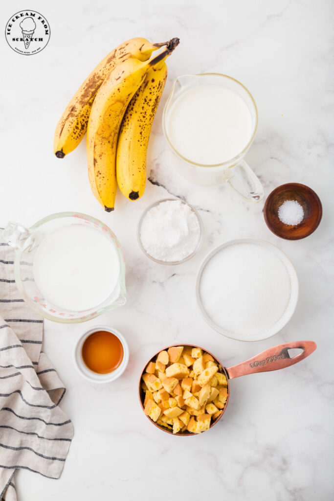 Ingredients needed for banana pudding ice cream on a marble countertop, all in separate bowls. Includes ripe bananas, chopped cookies, sugar, and cream.