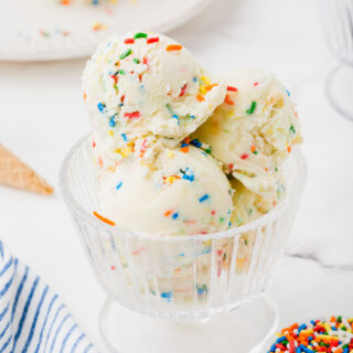 a clear glass ice cream dish filled with scoops of birthday cake ice cream with sprinkles. a small bowl of sprinkles is in the foreground.