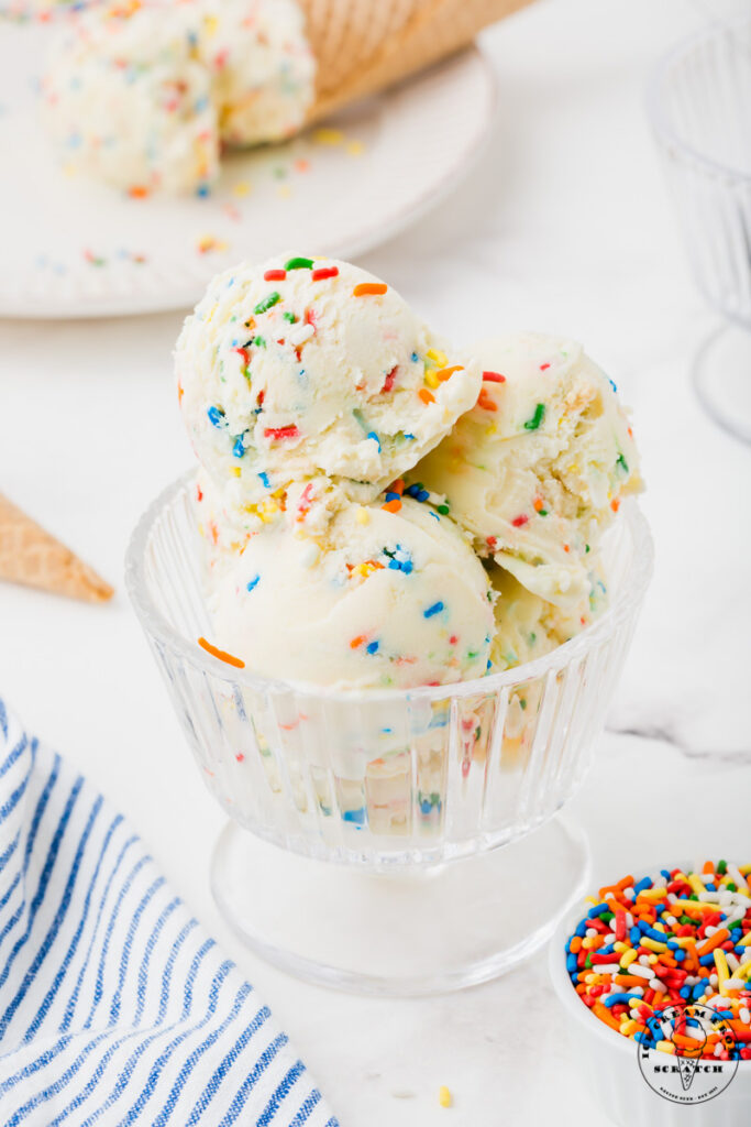 a clear glass ice cream dish filled with scoops of birthday cake ice cream with sprinkles. a small bowl of sprinkles is in the foreground.