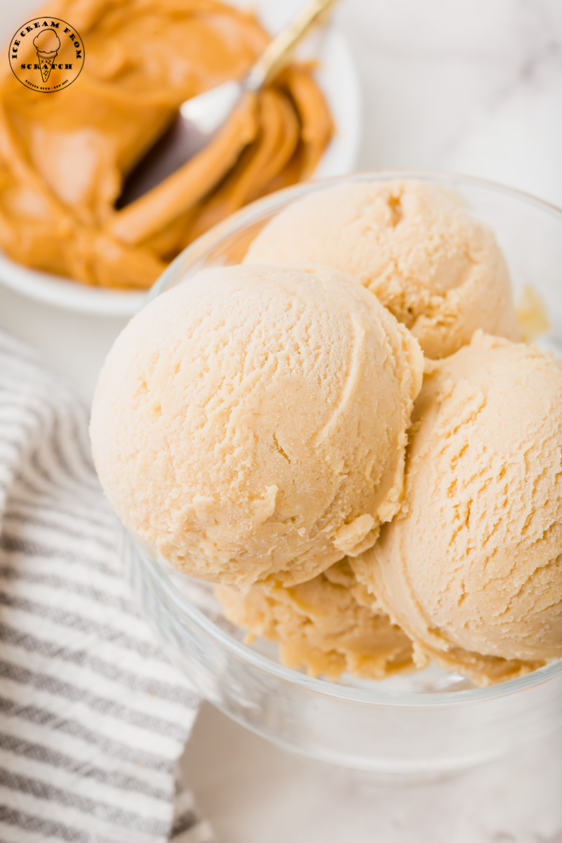 close up view of a glass bowl filled with scoops of creamy peanut butter ice cream. A bowl of peanut butter is in the background.