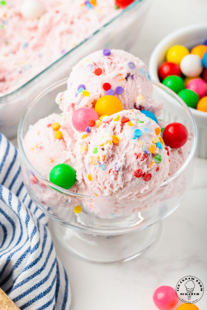 A glass dish of pink bubblegum ice cream with rainbow sprinkles and garnished with gum balls.