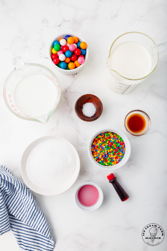 Ingredients for making bubblegum ice cream on a marble countertop. Includes sprinkles, cream, and gumballs
