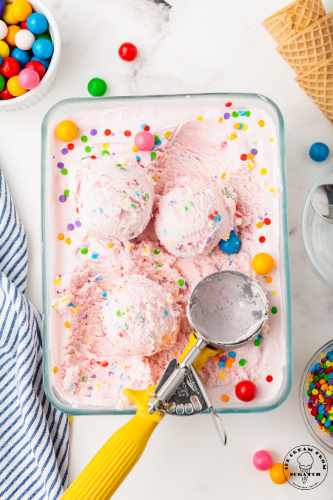 a glass baking dish filled with pink bubblegum ice cream. A yellow and metal ice cream scoop is making scopps, and the counter is filled with cones and colorful sprinkles.
