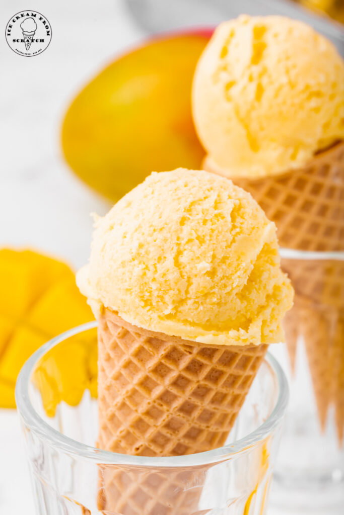 Two glasses holding up sugar cones topped with mango ice cream. Sliced mangoes are in the background
