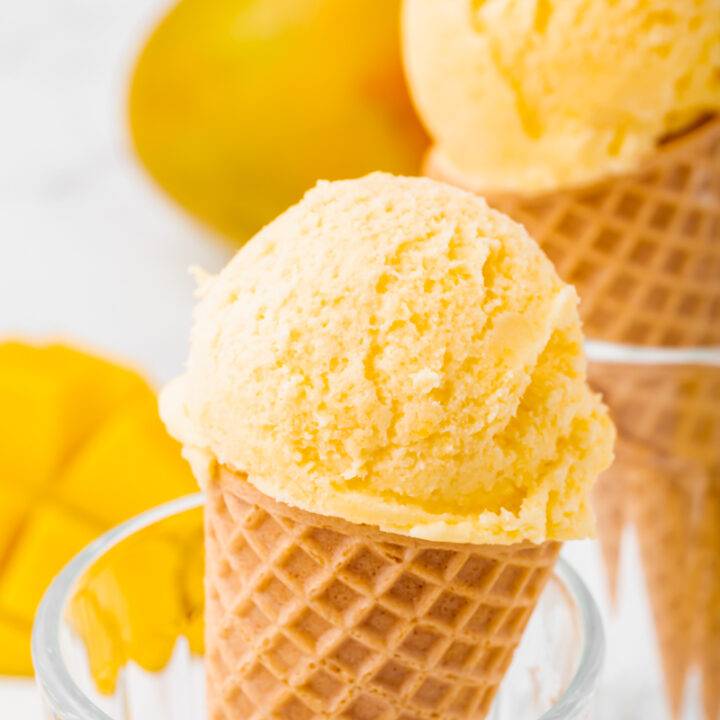 Two glasses holding up sugar cones topped with mango ice cream. Sliced mangoes are in the background