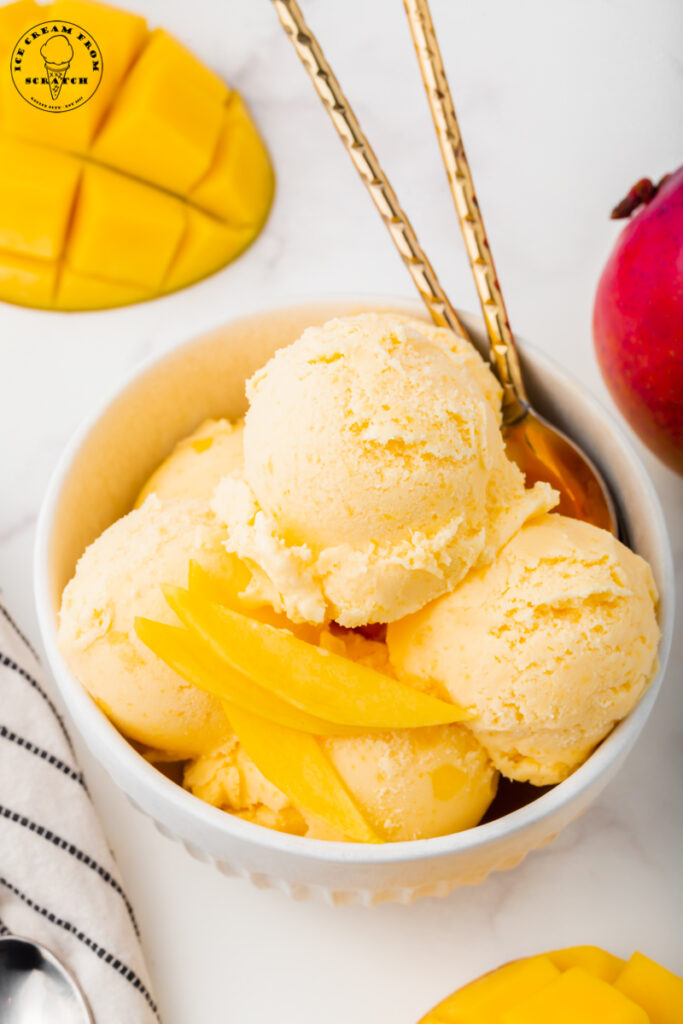 a large bowl of scooped mango ice cream topped with thinly sliced fresh mangoes. There are two gold spoons in the bowl.
