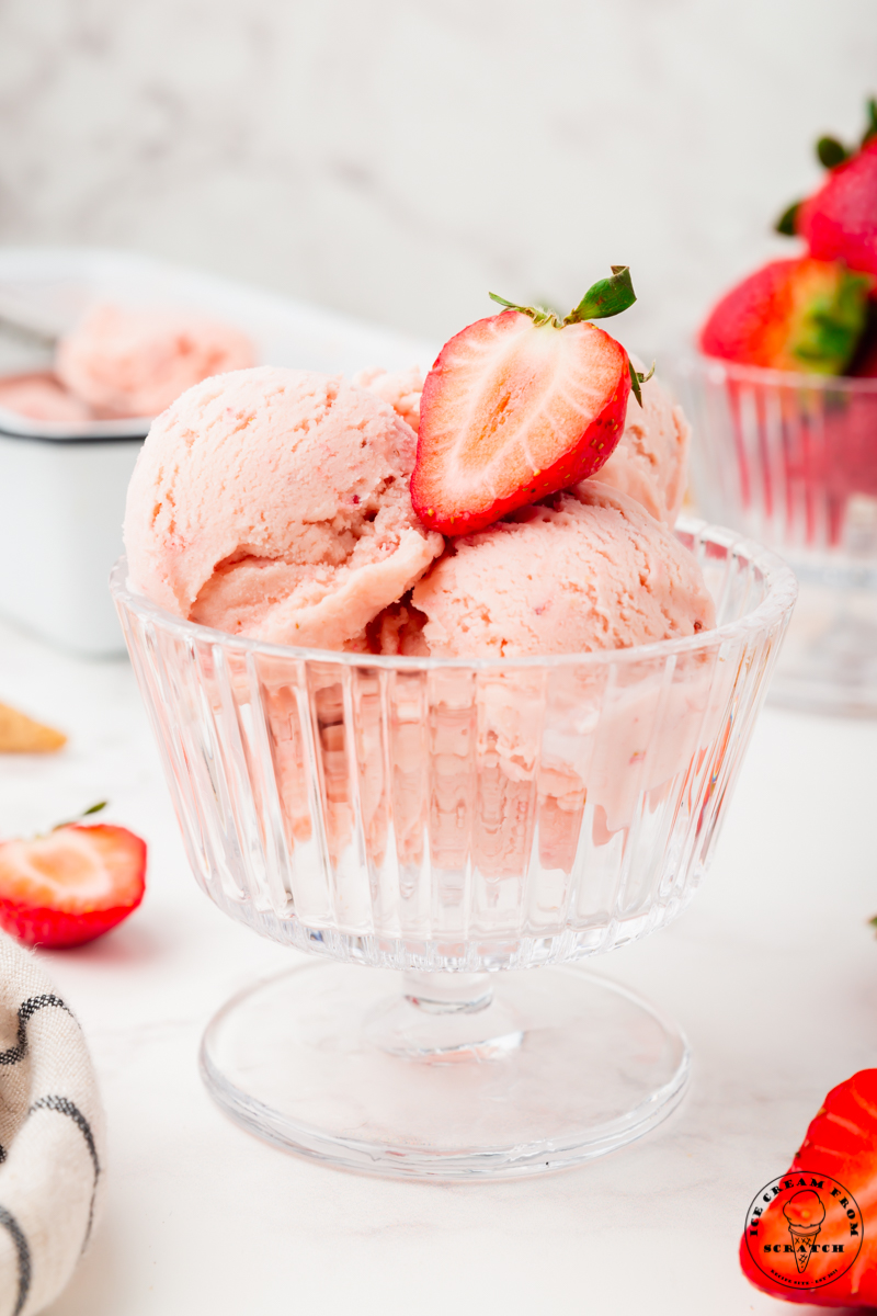 a footed glass dessert dish is filled with homemade strawberry ice cream and topped with half of a fresh strawberry.