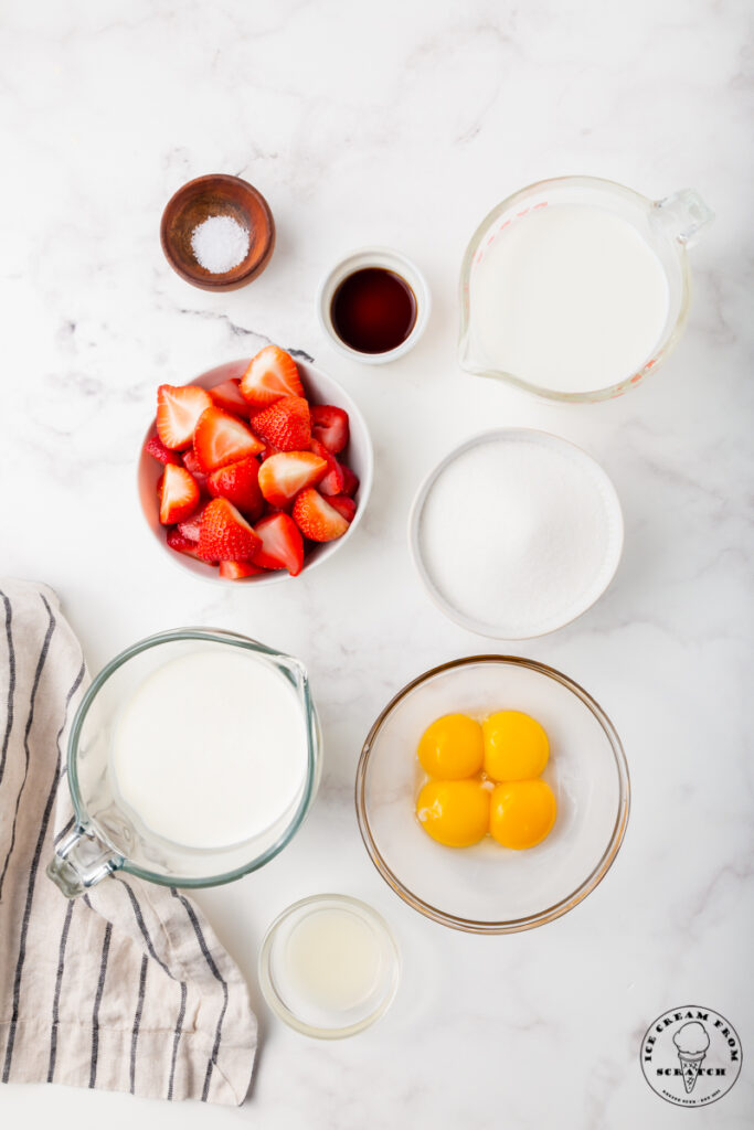 Ingredients for homemade strawberry ice cream are in separate bowls on a marble counter. Includes fresh berries, egg yolks, sugar and cream.