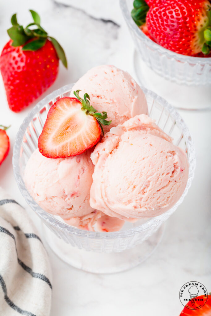 a footed glass dessert dish is filled with homemade strawberry ice cream and topped with half of a fresh strawberry.