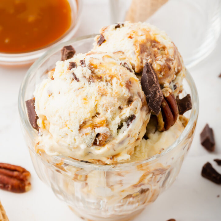 a glass dessert dish filled with scoops of turtle ice cream and garnished with pecans and chocolate chunks.