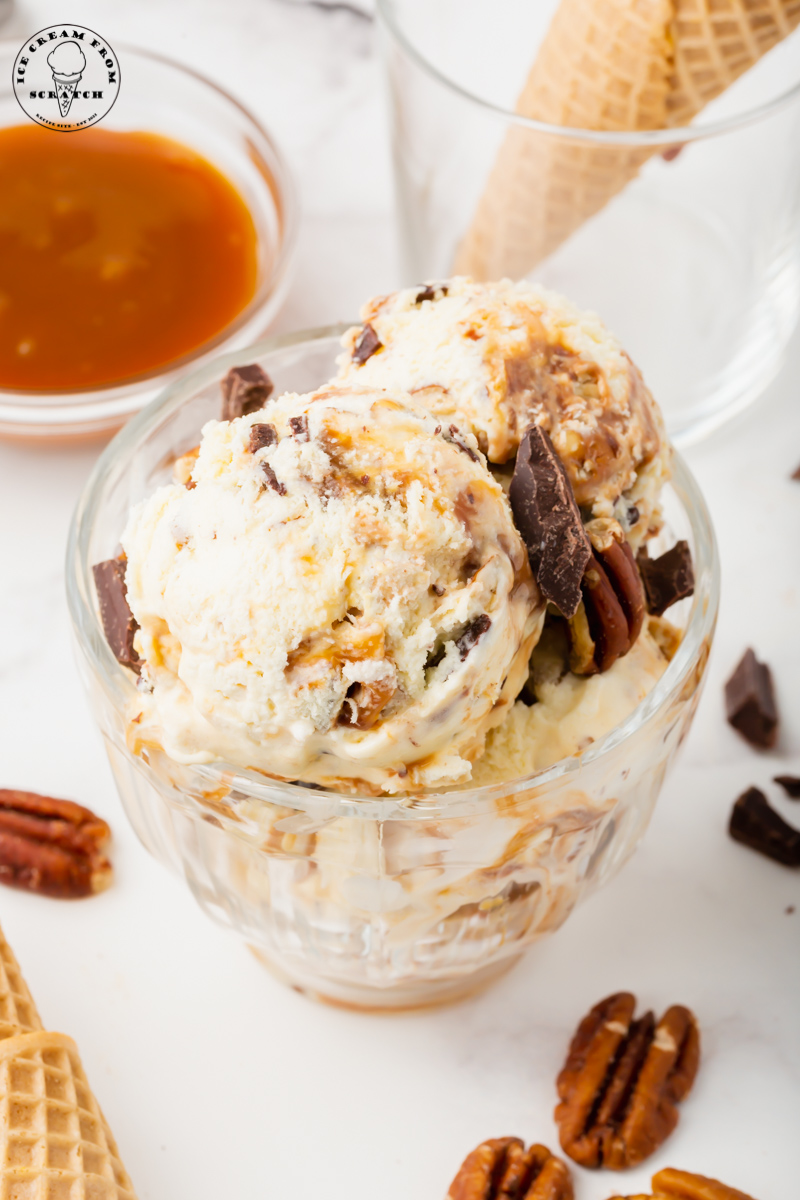 a glass dessert dish filled with scoops of turtle ice cream and garnished with pecans and chocolate chunks.