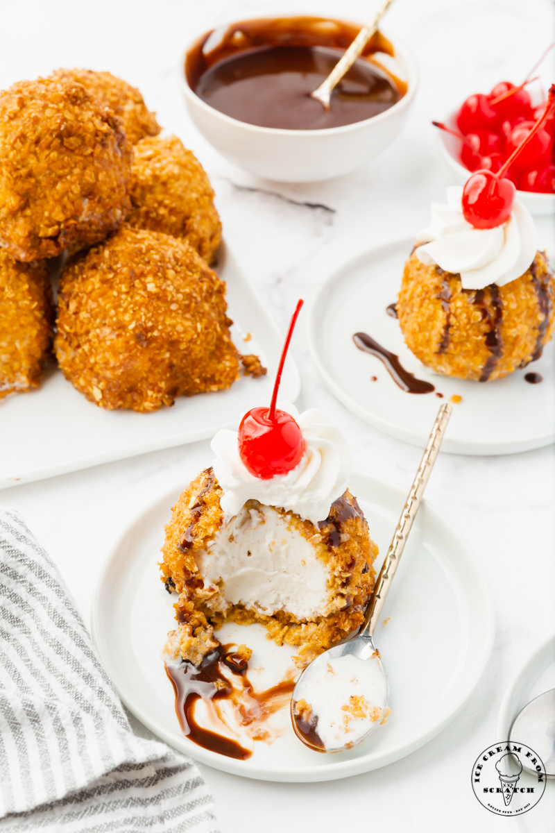 a table set with two servings of fried ice cream, one has been partially eaten. In the background is a bowl of chocolate syrup and a bowl of cherries