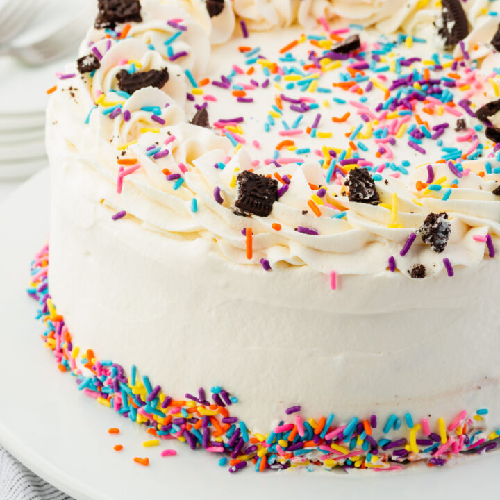 a round ice cream cake covered in frosting and topped with rainbow sprinkles and crushed Oreos.