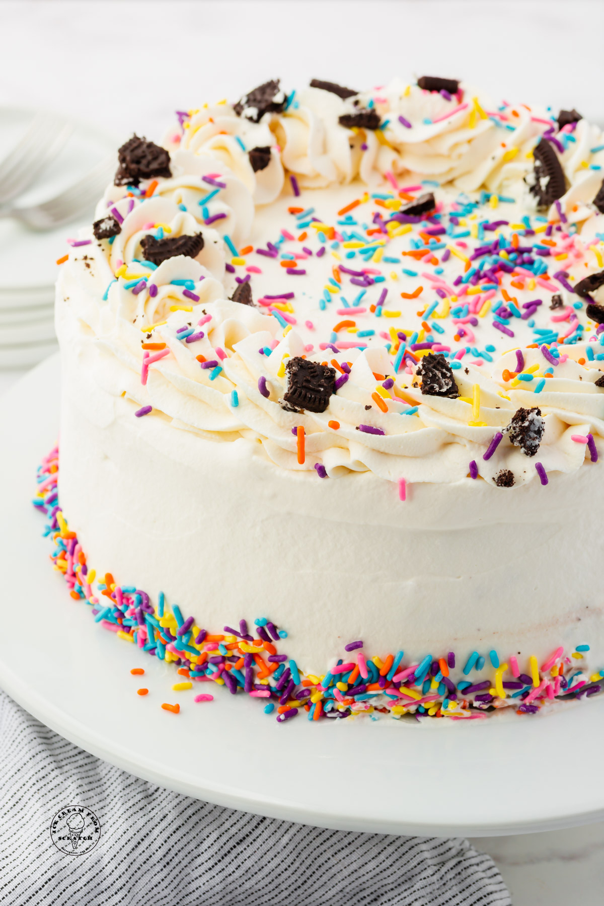 a round ice cream cake covered in frosting and topped with rainbow sprinkles and crushed Oreos.