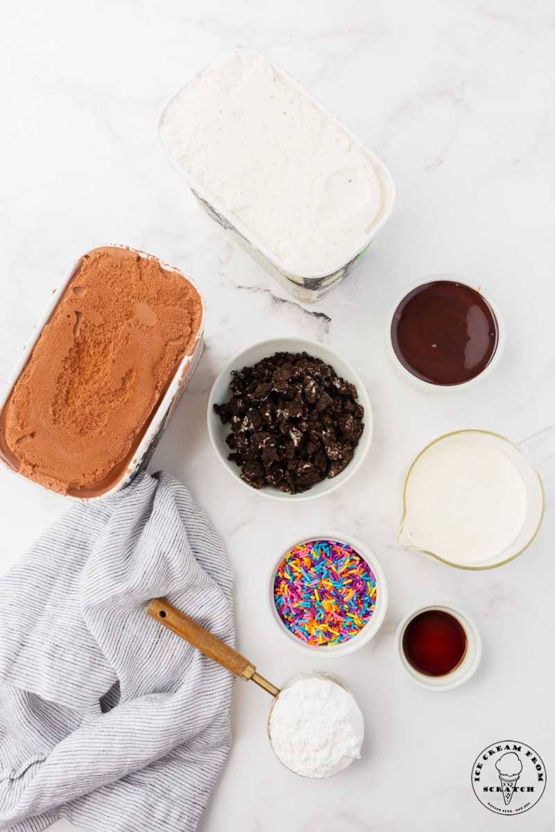 Ingredients for making a classic ice cream cake in separate bowls on a marble countertop. Includes both chocoalte and vanilla ice cream, crushed oreos, fudge sauce, and sprinkles.