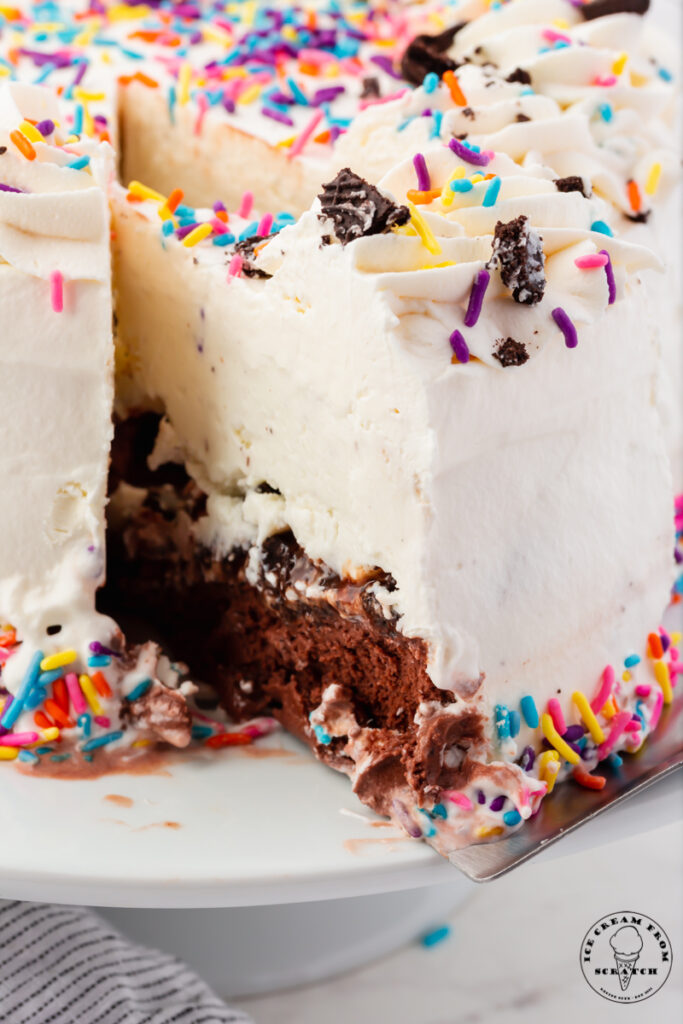 a slice of ice cream cake being pulled away from the whole cake with a cake server. Inside the cake is a layer of chocolate ice cream, cookies, and vanilla ice cream.
