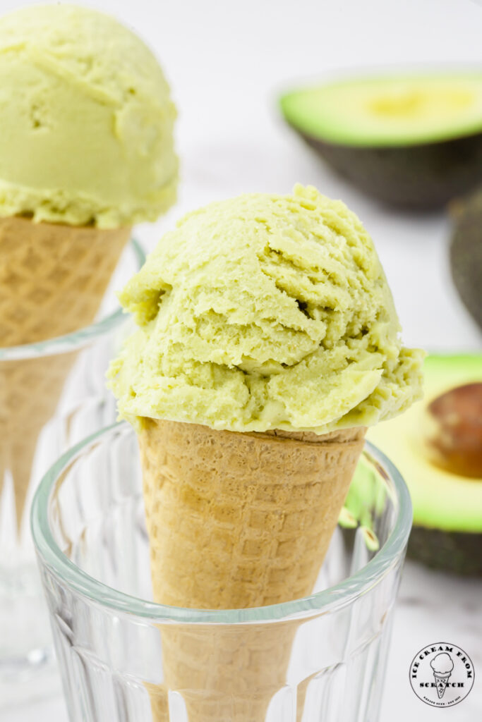 two sugar cones topped with green avocado ice cream, propped up in glasses. Halved avocados are in the background