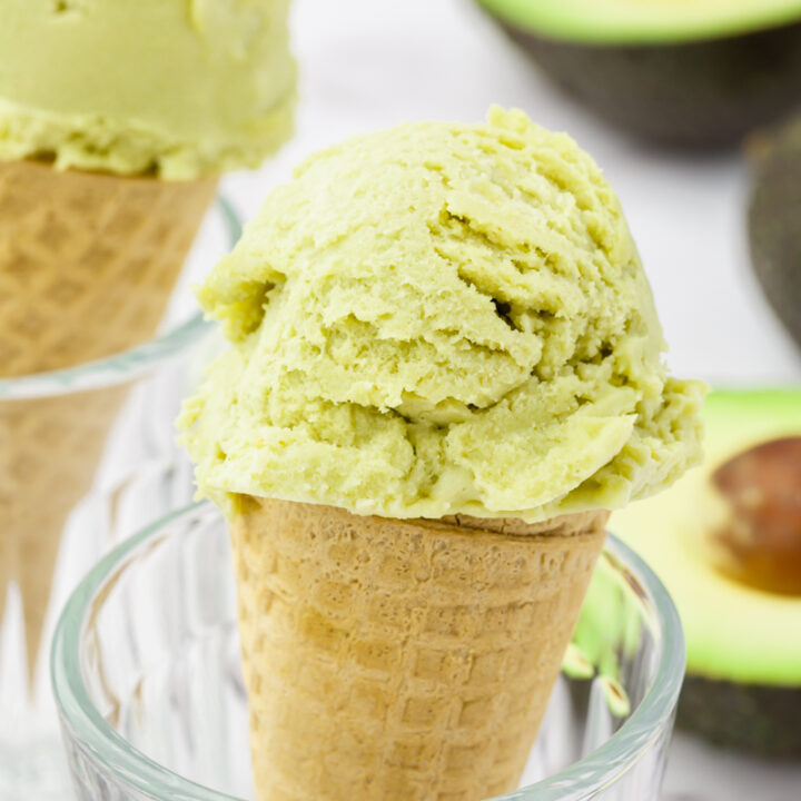 two sugar cones topped with green avocado ice cream, propped up in glasses. Halved avocados are in the background