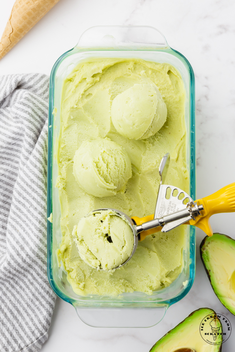 a glass dish of homemade avocado ice cream.  an ice cream scoop has created three scoops of it on top. View from above.