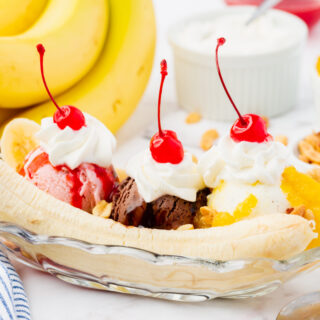 a long, shallow glass dish filled with a classic banana split topped with whipped cream and cherries. A bunch of ripe bananas is in the background.