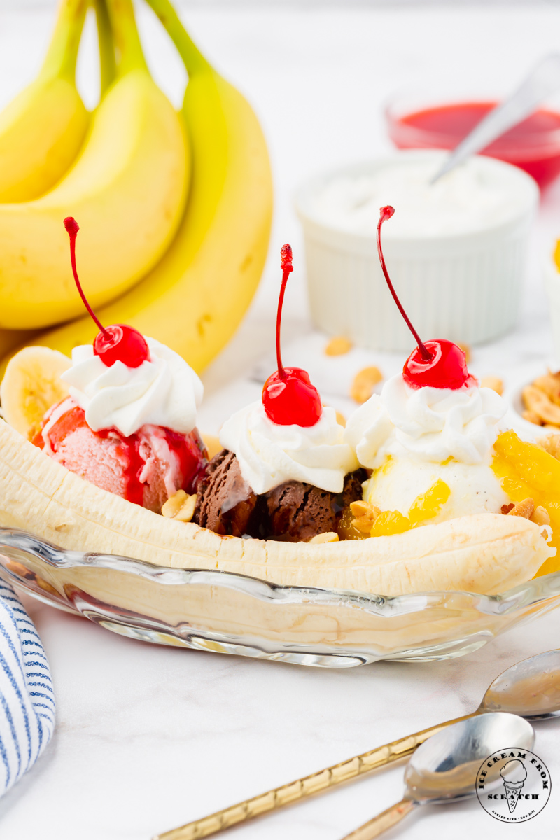 a long, shallow glass dish filled with a classic banana split topped with whipped cream and cherries. A bunch of ripe bananas is in the background. 
