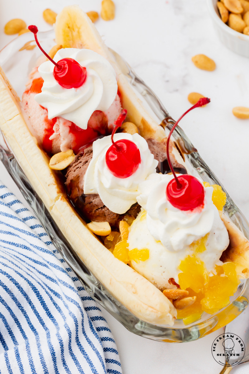 top view of a large banana split in a glass dish. Three scoops of ice cream are topped with various toppings, whipped cream, peanuts, and maraschino cherries.